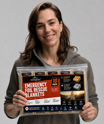 A smiling woman holds a clear plastic bag containing ten packaged emergency foil rescue blankets against a plain light gray studio background. The front label reads 'EMERGENCY FOIL RESCUE BLANKETS' and '10 PACK,' with the brand Mountain Gear visible near the top.