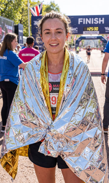 A smiling female marathon runner standing in front of a finish line arch, wrapped in a reflective silver and gold foil emergency blanket. She is wearing a race bib and black shorts, with volunteers and other runners visible in the background.