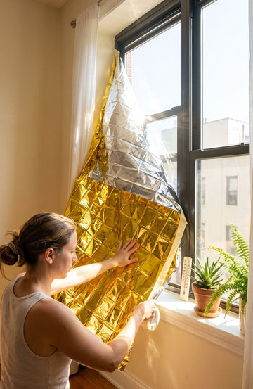 A woman in a white tank top is applying reflective gold foil insulation to a window in a bright room. The window has black frames, and a small potted plant sits on the sill.