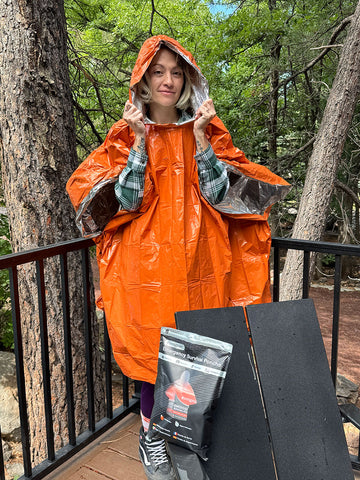 A woman wearing an orange waterproof poncho stands on an outdoor wooden deck surrounded by trees, holding up the hood. A black package labeled 'Rain Gear' and two black panels are placed in front of her.