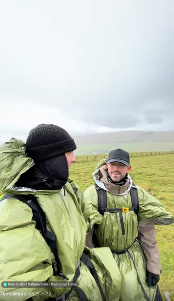 Two people wearing olive green waterproof parkas and winter gear standing outdoors in a grassy landscape under an overcast sky.