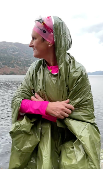 A woman wearing a shiny olive green rain poncho and a pink running headband stands outdoors by a lake with mountains in the background.