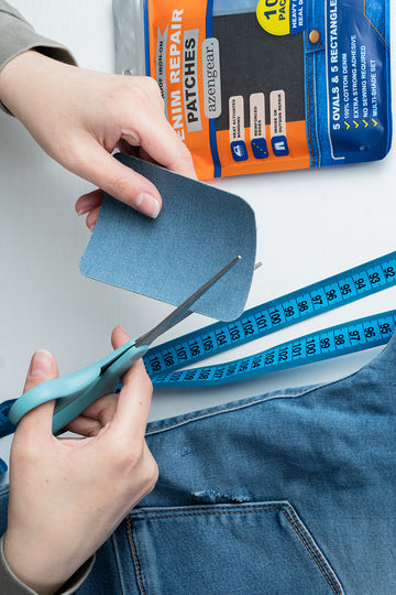 A person's hands are shown using blue-handled scissors to cut a piece of blue denim fabric, with a measuring tape and a package of denim repair patches visible in the background.
