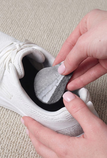 A close-up of a person's hands placing a gray, teardrop-shaped absorbent pad into the opening of a white athletic shoe on a beige carpeted floor.