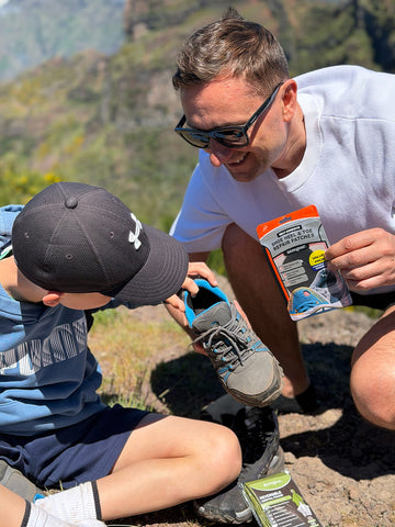 A man and a child outdoors on a mountain trail, with the man holding a packaged pair of gray trail running shoes and the child looking closely at the shoe. The man wears a white t-shirt, sunglasses, and a black Under Armour cap, while the child wears a blue shirt and dark shorts.