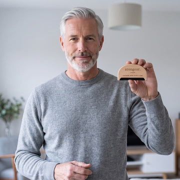 An older man with gray hair and a beard wearing a gray sweater, holding up a small wooden automatic pet feeder bowl in his right hand. The feeder is light wood with black text that reads 'azenzear.