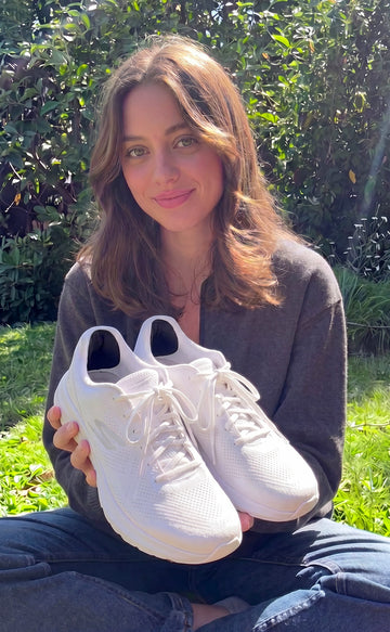 A woman sitting outdoors on grass in front of green bushes, holding a pair of white athletic sneakers.